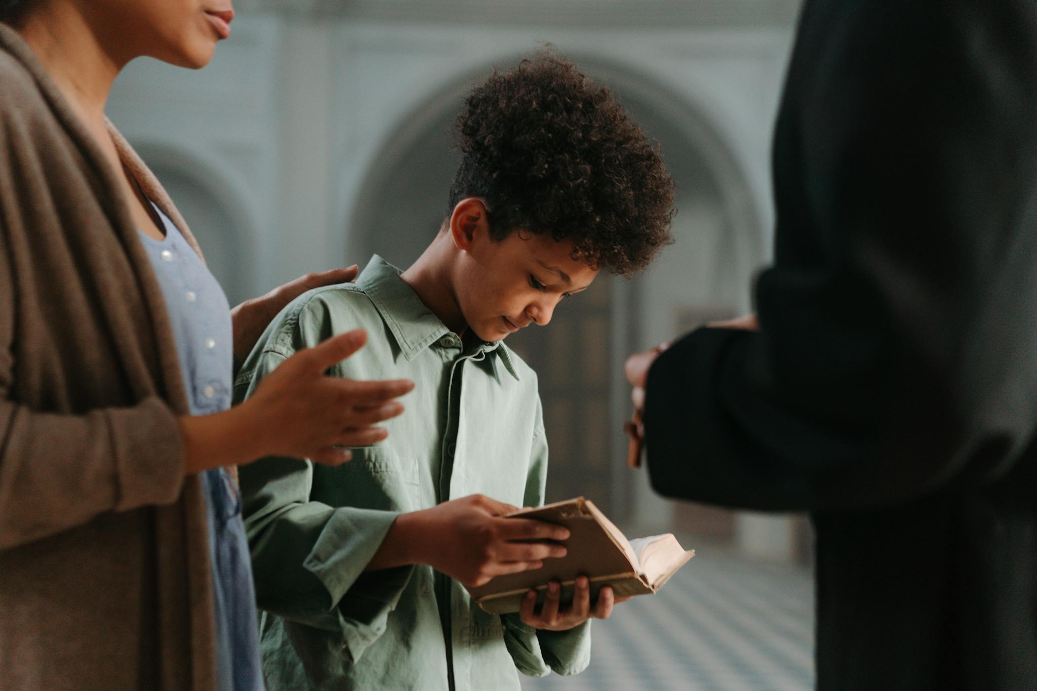 A young boy reads a religious book with guidance from adults in an indoor church setting.